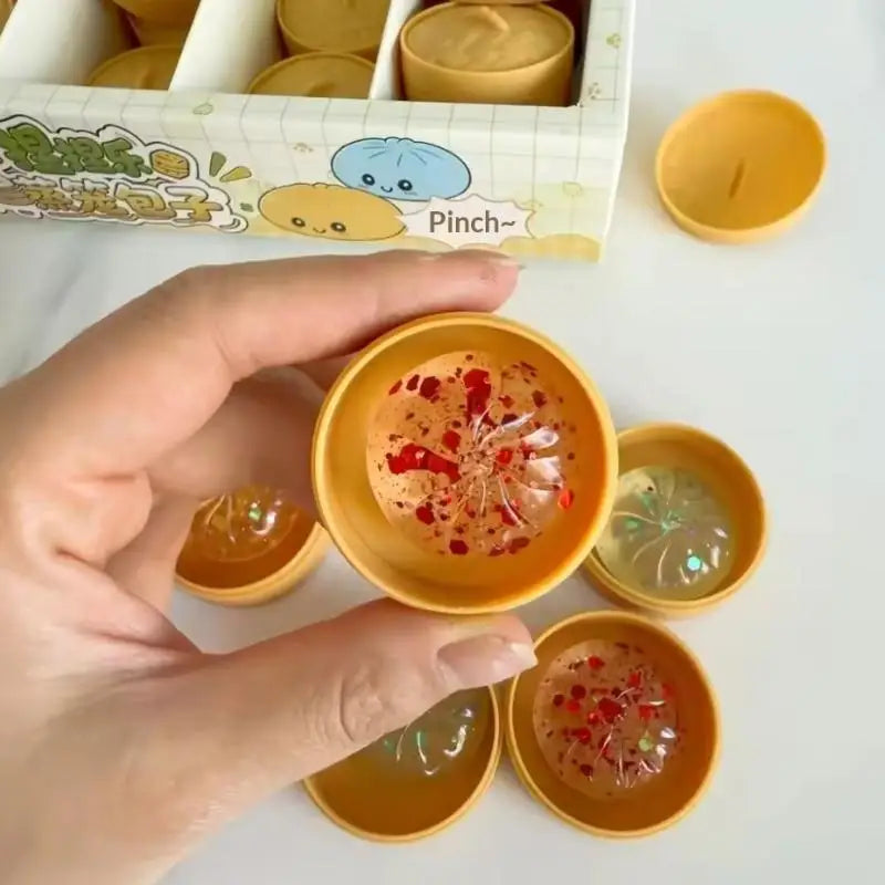 A hand holding a mini soup dumpling squishy in its bamboo steamer case, with more coloured dumpling squishies below.